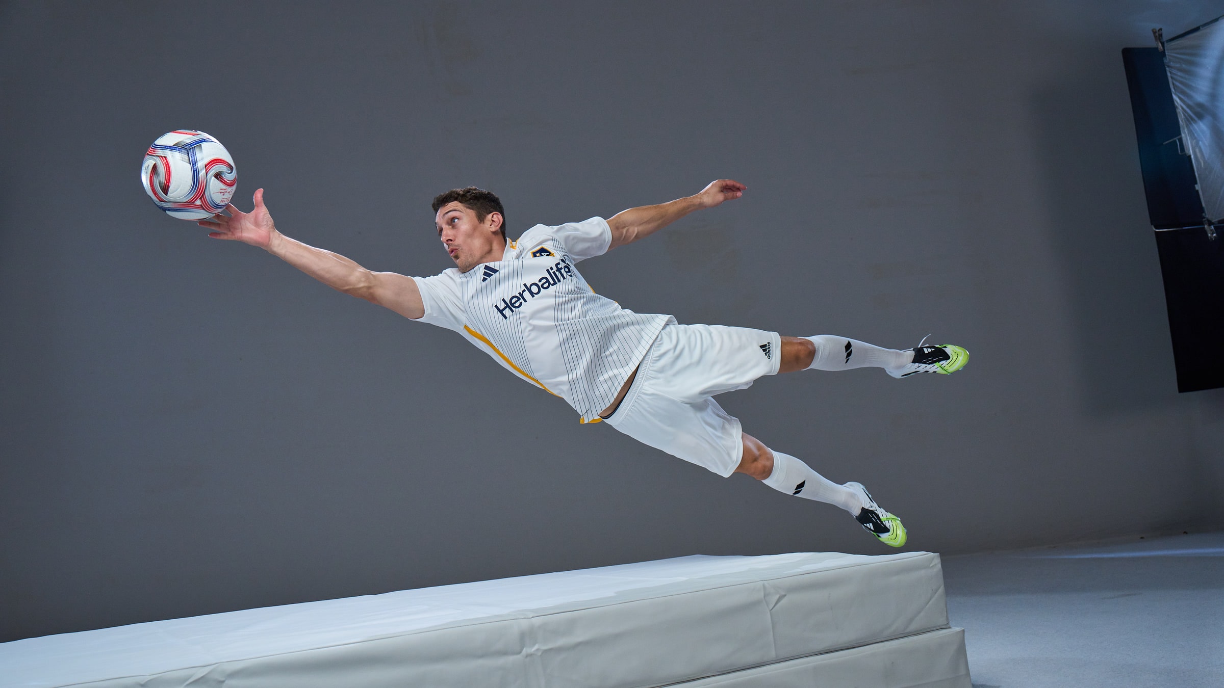 Bryan Jordan at full stretch in mid-air in an LA Galaxy kit, reaching for the ball on a studio cyclorama with crash mats below.
