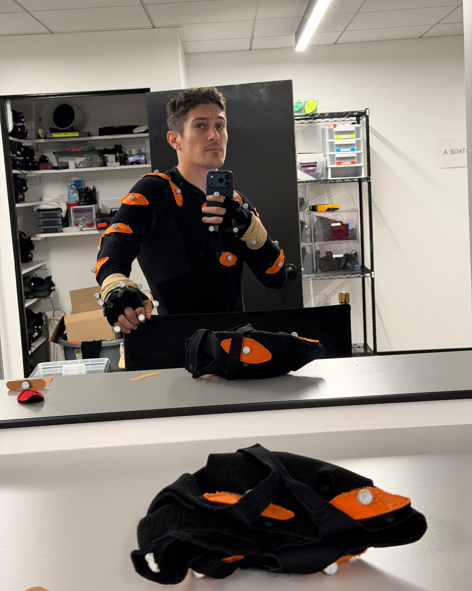 Bryan Jordan in a motion capture suit with orange marker pads, photographing himself in a mirror at the marker station, spare suit pieces on the counter.