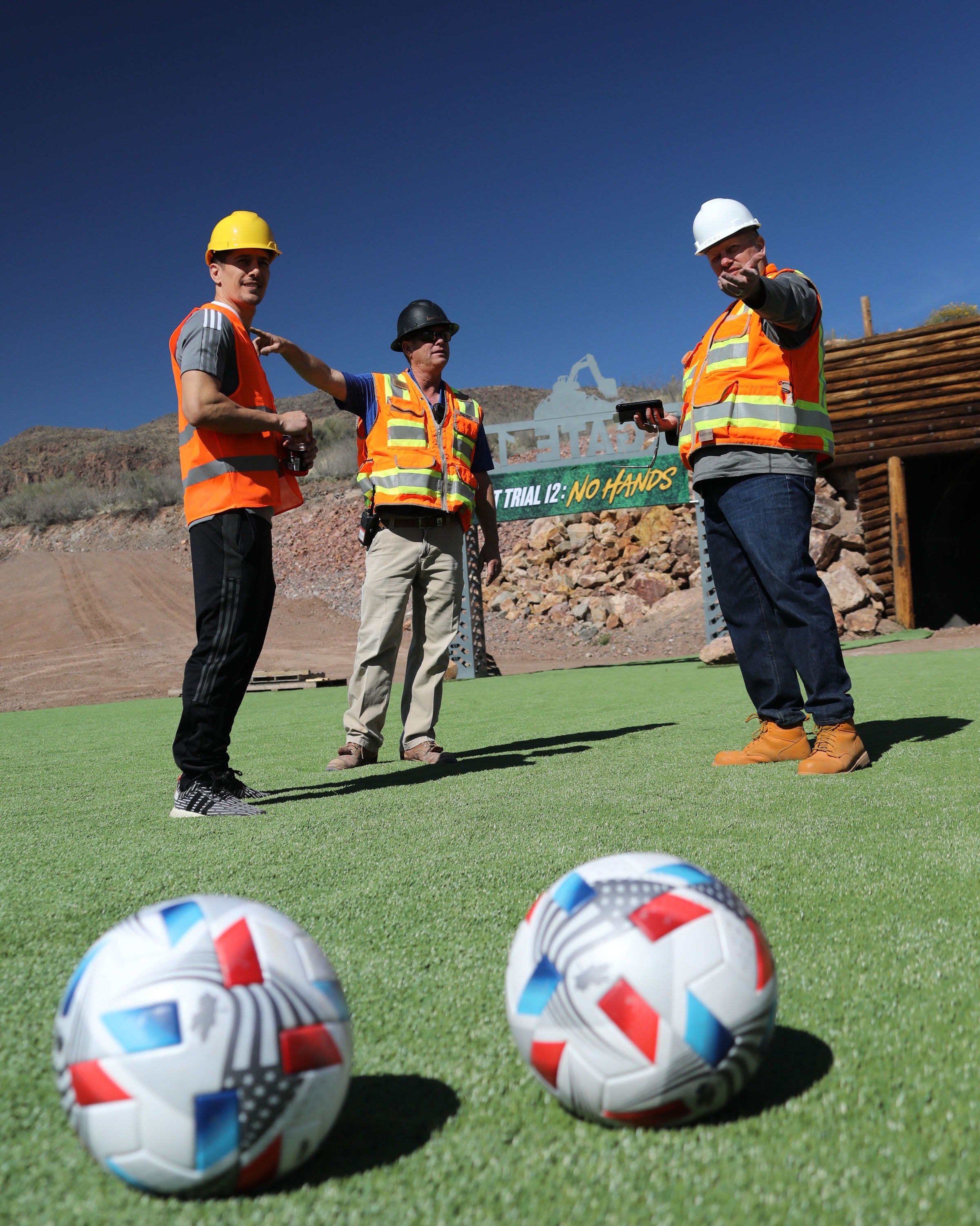 Three people in hard hats and hi-vis vests on a turf pitch at the Cat Trial 12 site, two match balls in the foreground.