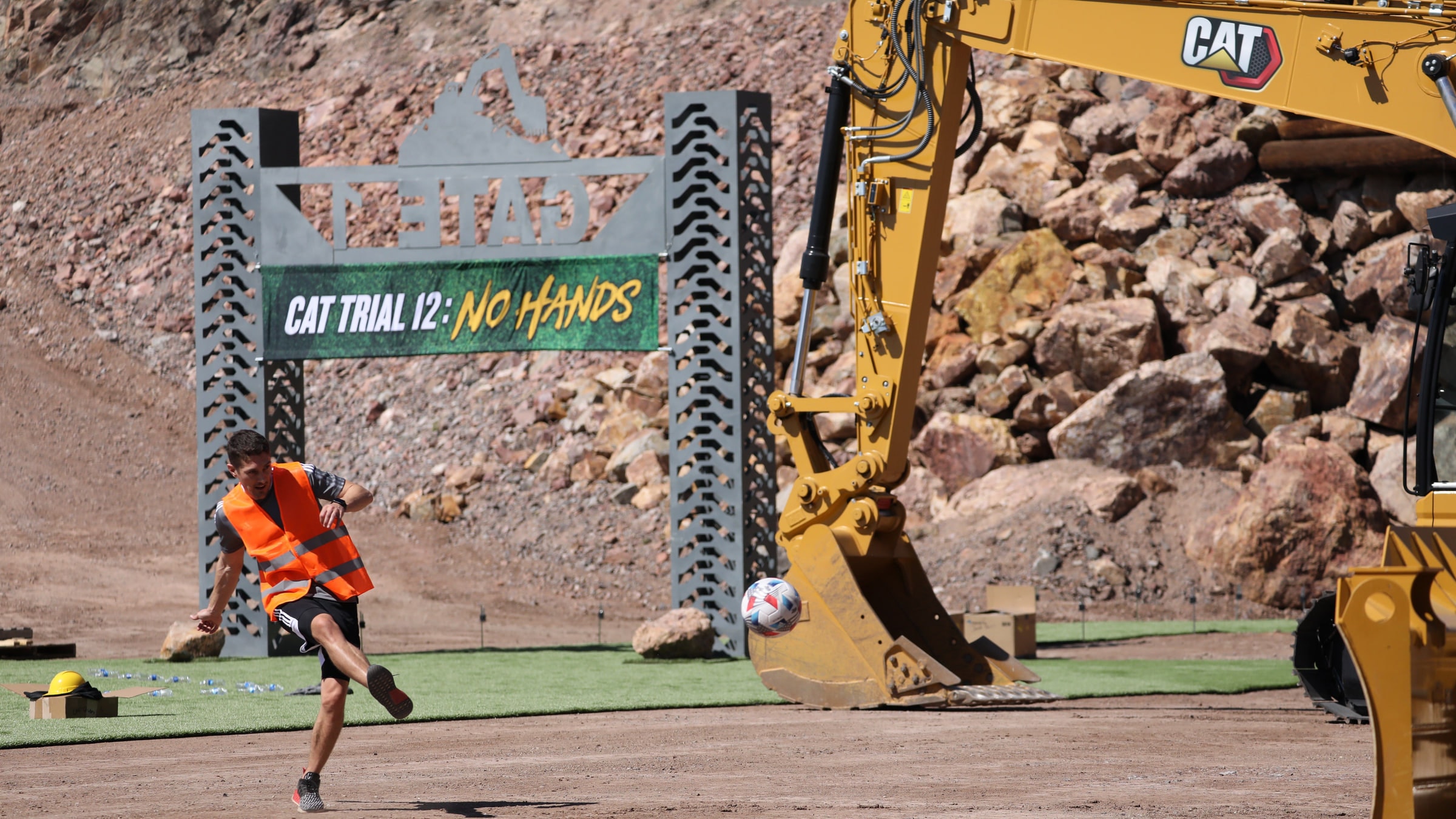 Bryan Jordan in a hi-vis vest striking a ball toward an excavator bucket, the Cat Trial 12 start gate behind him.