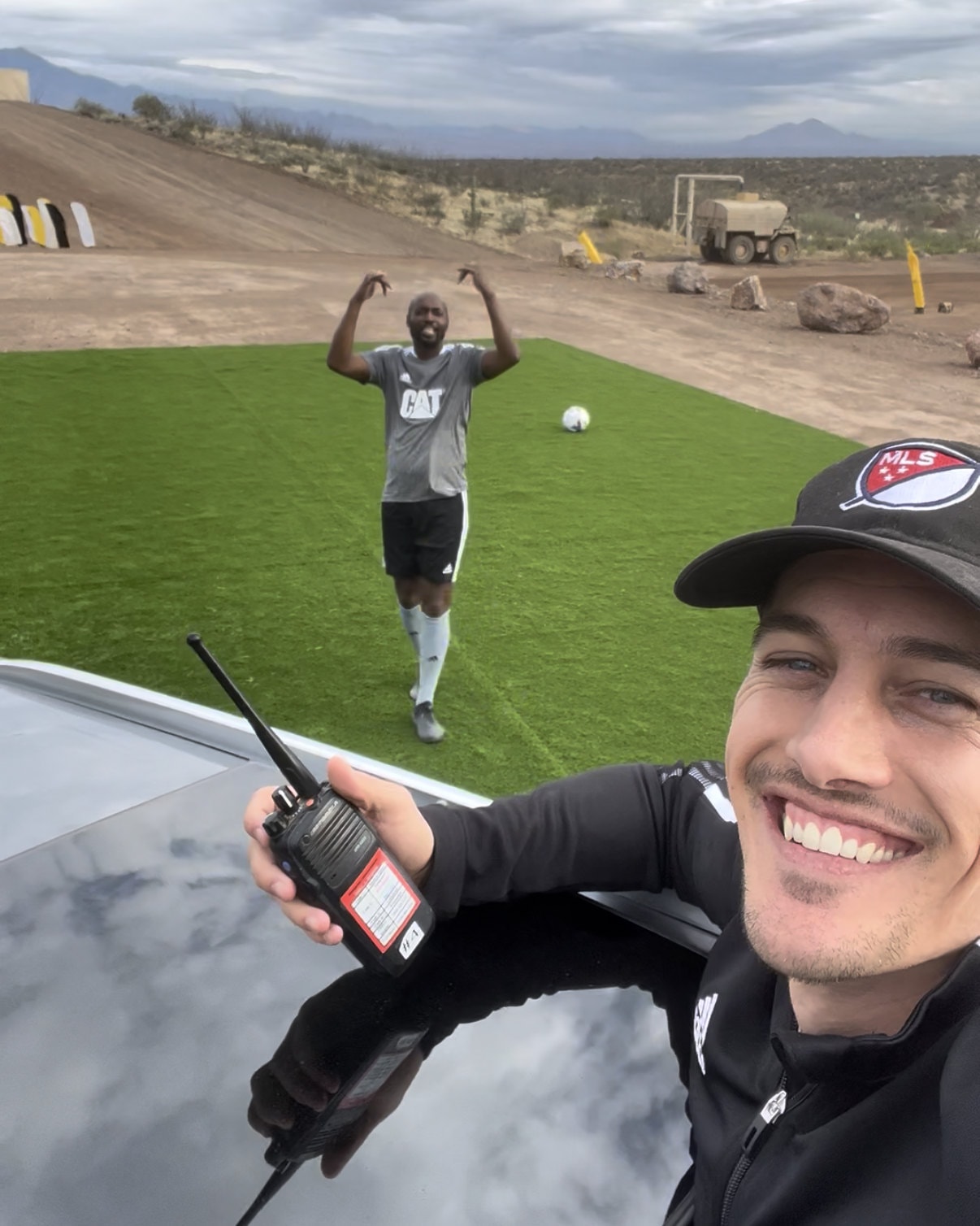 Bryan Jordan holding a radio on the Cat Trial 12 set, DaMarcus Beasley on the turf pitch behind him.